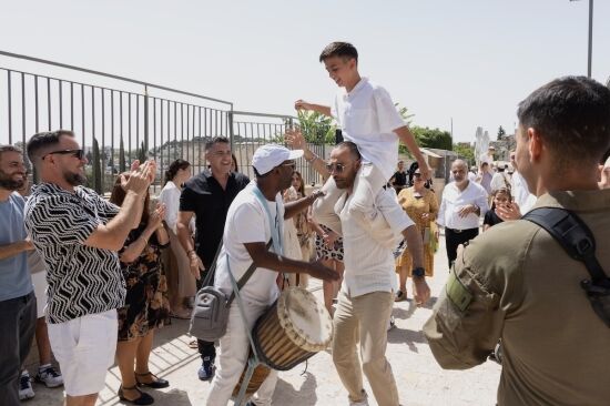תהלוכת בר מצווה בכותל – נער בר מצווה מורם על הכתפיים, מתופף מרים את הקהל בשירה וריקודים, משפחה ואורחים מוחאים כפיים בשמחה