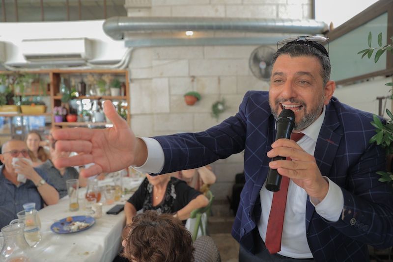 פייטן בחליפה כחולה עם עניבה אדומה מחייך לאורחים תוך כדי שירה במיקרופון באירוע משפחתי.