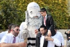 משפחה וחברים חוגגים עם בובת ענק בדמות רב בחוץ, עם רקע של עצים וילד במדים מחויך שנוגע בבובה.